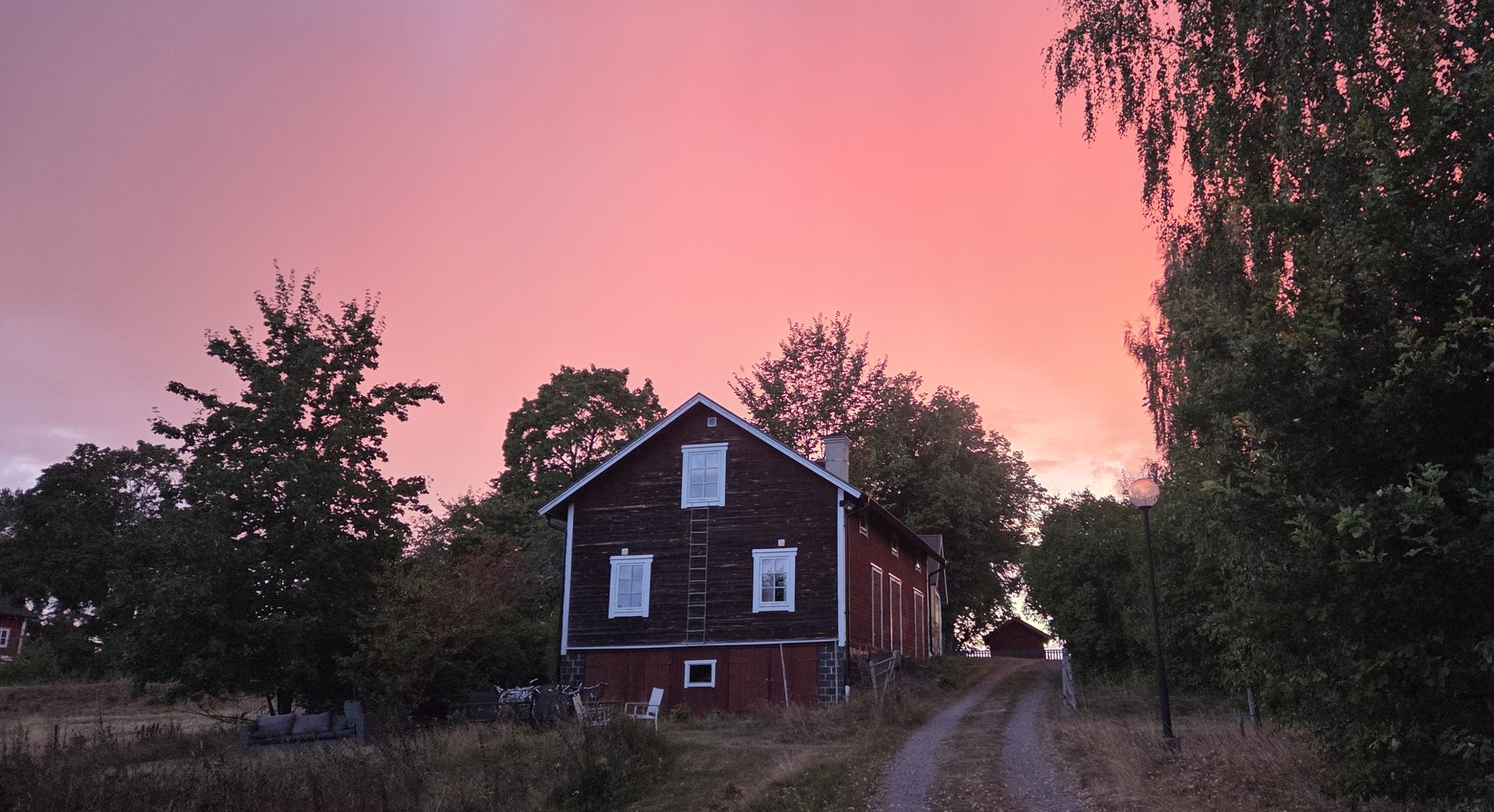 Gården i Skedevi i solnedgång, med grusvägen fram mot det röda timmerhuset och himlen i korallrött.