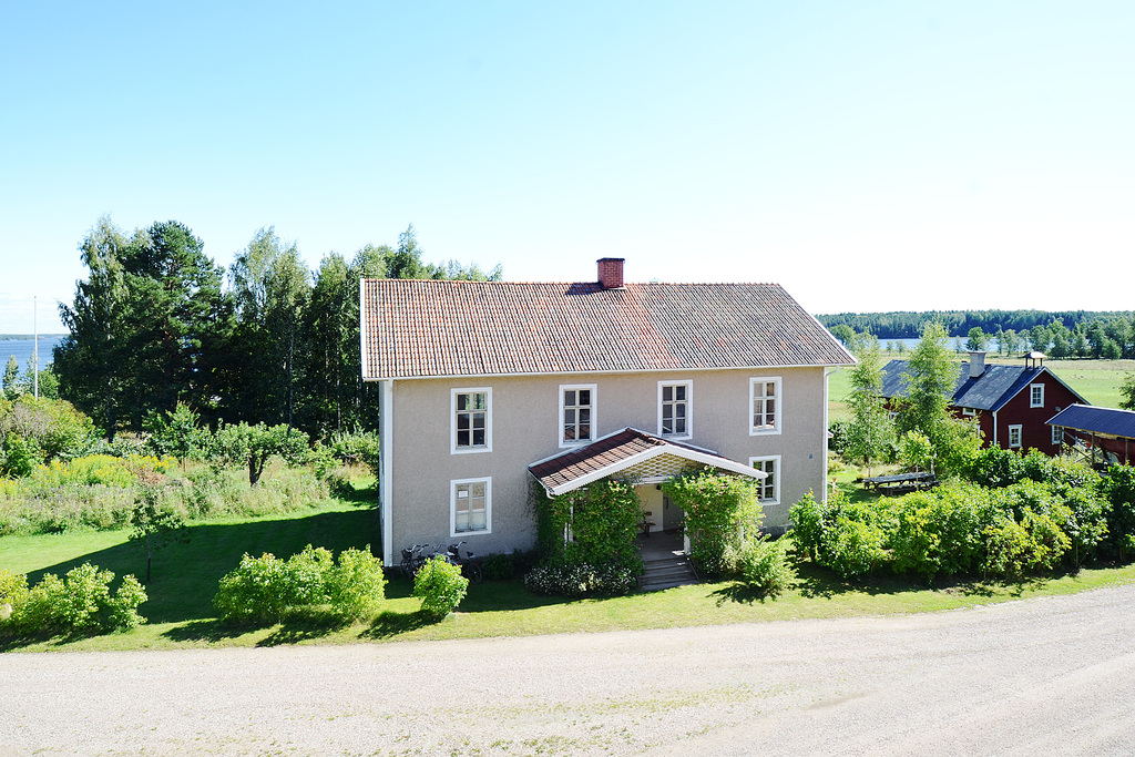 Vitputsat hus med veranda och trädgård, sjön skymtar bakom.