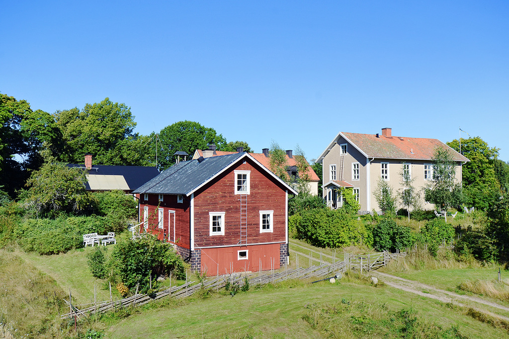 Röd timmerbyggnad och putsat hus med gärdesgård framför.